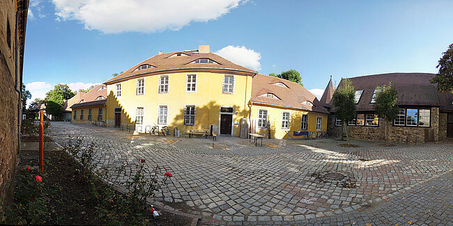 Mensa Burg; Foto: Thomas Faust Ein sonniger Platz mit Kopfsteinpflaster, umgeben von historischen Gebäuden. Im Vordergrund befinden sich Blumen und ein Brunnen. Die Gebäude haben gelbe und braune Fassaden mit weißen Fenstern. Der Himmel ist blau mit einigen Wolken. Die Atmosphäre wirkt ruhig und malerisch.