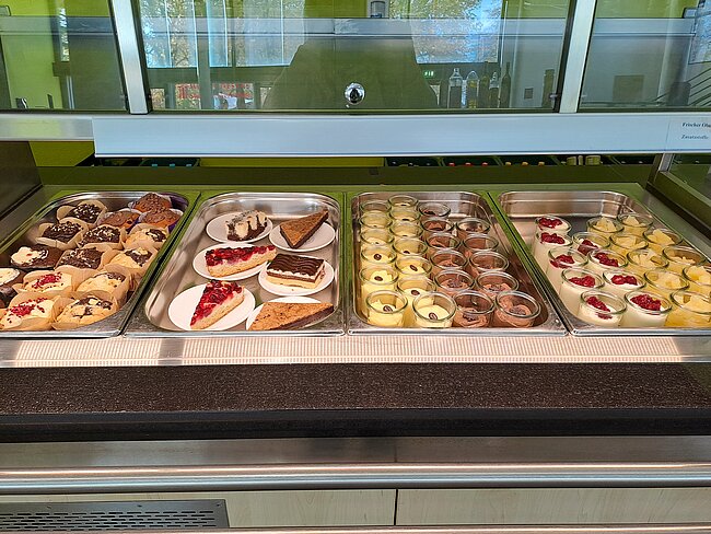 Ein Buffet mit verschiedenen Desserts, darunter Kuchenstücke, Donuts, kleine Törtchen und andere Süßspeisen. Die Speisen sind in durchsichtigen Behältern auf einem langen Tisch angeordnet.