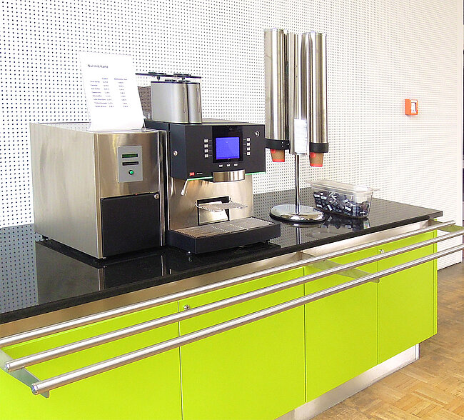 Kaffeeautoimat Mensa Bernburg; Foto: Thomas Faust Ein modernes Kaffeeautomatensystem auf einer grünen Arbeitsplatte. Der Kaffeeautomat hat einen Bildschirm und verschiedene Ausgabestationen für Kaffee und Milch. Daneben steht ein Glas mit Kaffeelöffeln.