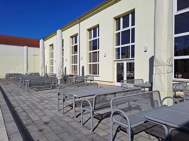 "Ein Außenbereich mit mehreren Tischen und Stühlen, die auf einem gepflasterten Boden aufgestellt sind. Die Tische und Stühle sind aus Metall und stehen vor einem hellgelben Gebäude mit großen Fenstern. Der Himmel ist klar und blau."
