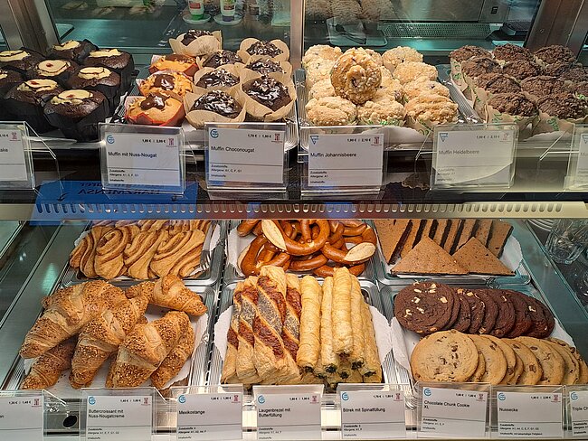 Ein Bild eines gut sortierten Bäckereiregal mit verschiedenen Süßgebäcken. Oben befinden sich Kuchen wie Schokoladenkuchen, Obstkuchen und Biskuitrollen. Unten sind Croissants, Zimtschnecken, Blätterteiggebäck, Kekse und andere Backwaren. Jedes Produkt hat ein Schild mit dem Namen und Preis.