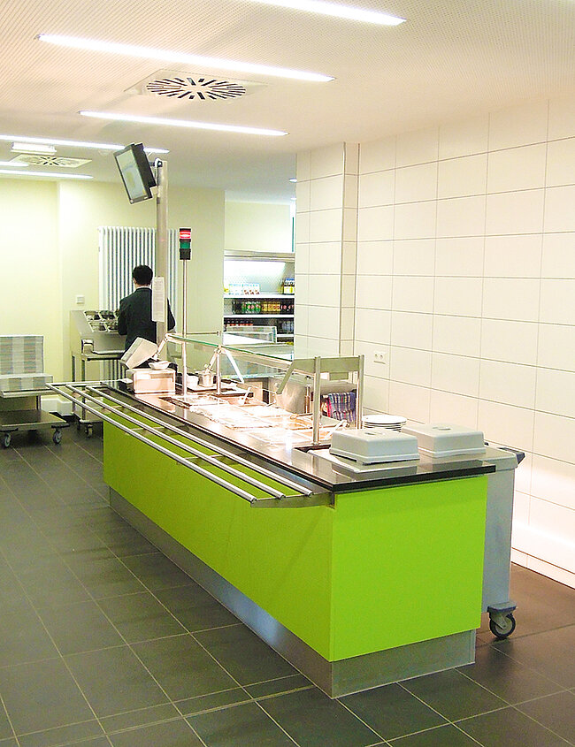 Speisenausgabe Mensa Bernburg; Foto: Thomas Faust Ein sauberer, moderner Verkaufstresen in einem Lebensmittelgeschäft oder einer Bäckerei. Der Tresen ist grün und hat eine gläserne Abdeckung. Im Hintergrund ist eine Person zu sehen, die in der Küche arbeitet. Die Wände sind weiß gefliest.