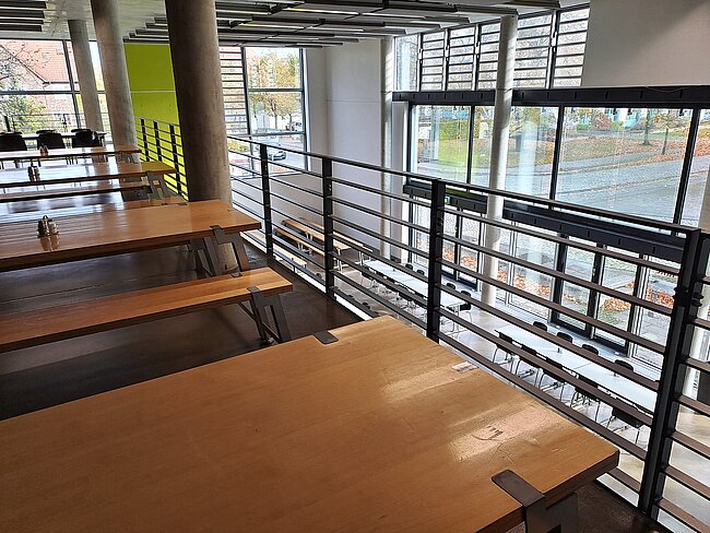 "Ein moderner Innenraum mit langen Holztischen und Stühlen. Große Fenster bieten einen Blick auf die Straße draußen. Der Raum ist hell und hat eine offene, luftige Atmosphäre mit Metallgeländern und Betonsäulen."