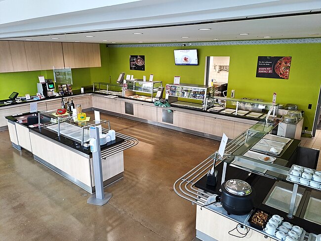 Ein sauberer, moderner Selbstbedienungsbereich in einem Restaurant oder einer Cafeteria. Es gibt verschiedene Theken mit Glastheken, in denen verschiedene Speisen und Getränke ausgestellt sind. Die Wände sind grün gestrichen, und es gibt einige Schilder und Anzeigen an den Wänden.