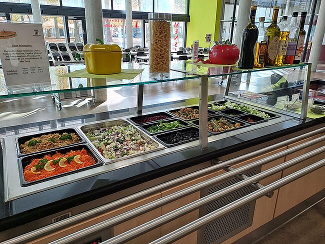 Ein Buffet mit verschiedenen Salaten in durchsichtigen Behältern. Dazu gehören Gemüsesalate, Nudelsalate und andere bunte Salatmischungen. Auf dem Buffet stehen auch große Gewürzflaschen und ein Schild mit Informationen. Der Bereich ist gut beleuchtet und wirkt einladend.
