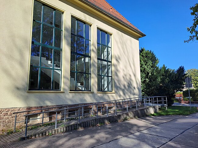 Ein Gebäude mit großen Fenstern und einer Rampe für Rollstuhlfahrer. Die Fassade ist hell und es gibt einen gepflasterten Weg daneben. Im Hintergrund sind Bäume und ein klarer blauer Himmel zu sehen.