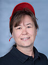 Eine Frau mit braunen Haaren trägt eine rote Baseballkappe und lächelt in die Kamera. Sie trägt ein schwarzes Polohemd. Der Hintergrund ist hellblau.