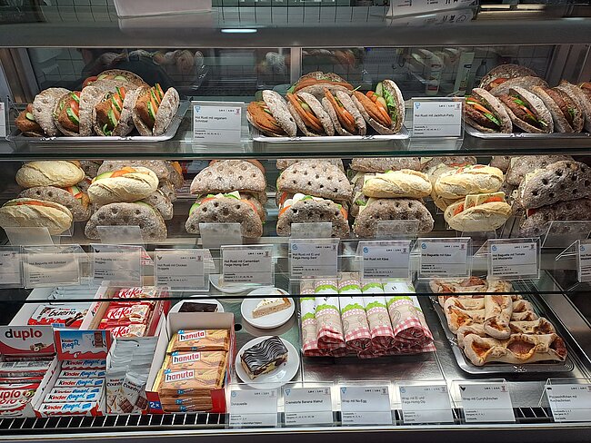 Ein Bild eines Supermarktregals mit verschiedenen Brotsorten und Backwaren. Unten befinden sich Aufschnitt, Käse und Snacks wie Duplo und Croissants. Die Produkte sind in Glastheken und Regalen ordentlich angeordnet und mit Preisschildern versehen.
