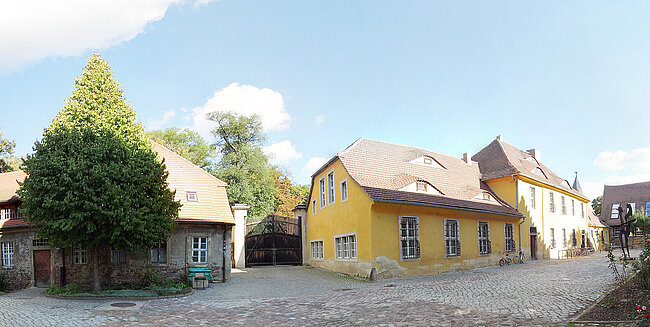 Mensa Burg; Foto: Thomas Faust Ein ruhiger, ländlicher Platz mit alten Gebäuden und Kopfsteinpflaster. Im Vordergrund steht ein großer Baum. Die Gebäude sind in verschiedenen Farben gestrichen, darunter ein gelbes Haus mit weißen Fenstern. Der Himmel ist blau mit einigen Wolken.
