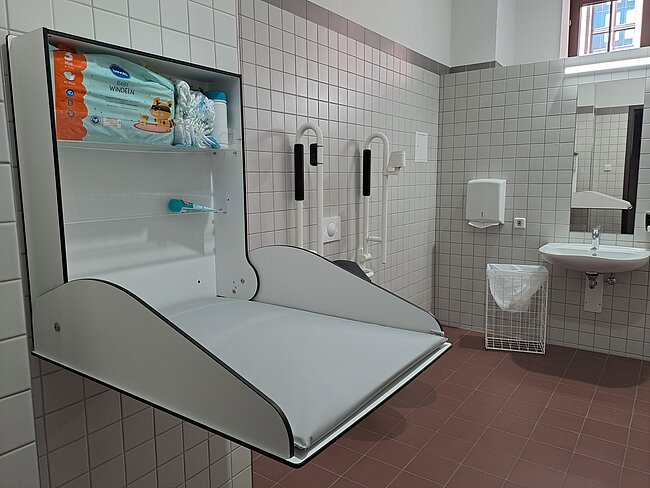 Ein behindertengerechter Waschraum mit einem speziellen Waschbecken, das für Rollstuhlfahrer geeignet ist. Es gibt Seifenspender, Handtuchhalter und einen Mülleimer. An der Wand sind Hygieneartikel wie Handschuhe und Bürsten angebracht. Im Hintergrund befindet sich ein weiterer Waschbereich mit einem Waschbecken und Spiegel.