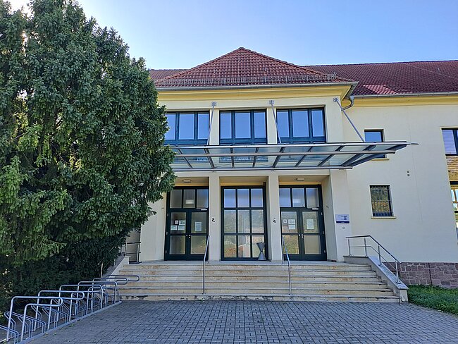 Ein Gebäude mit gelber Fassade, rotem Dach und mehreren großen Fenstern. Der Eingang besteht aus einer doppelten Glastür mit einem überdachten Vordach. Vor dem Gebäude befindet sich ein Baum und eine kleine Treppe mit einem Handlauf. Der Boden ist gepflastert.