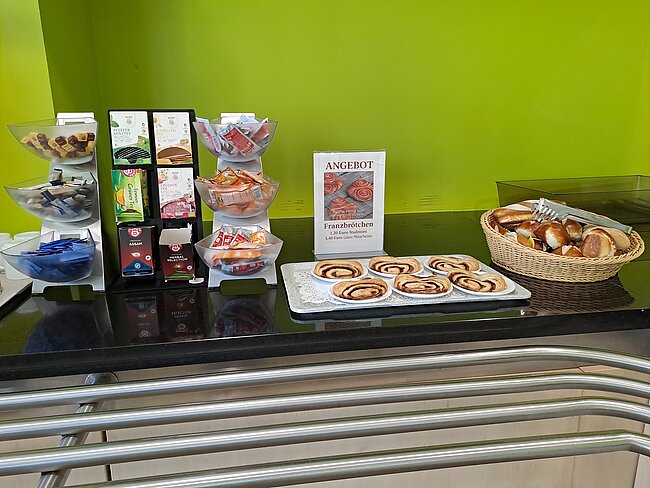 Ein Buffet mit verschiedenen Gebäcksorten, darunter Croissants, Zimtschnecken und andere Süßigkeiten. Die Auswahl ist auf einem Tisch mit einer Glasplatte präsentiert. Ein Schild mit der Aufschrift "ANGEBOT" und "Frühstücksbar" ist sichtbar.
