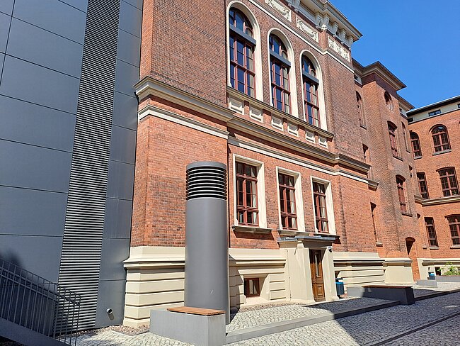 Die Mensa von außen Ein Backsteingebäude mit großen Fenstern und einem modernen grauen Metallrohr im Vordergrund. Der Himmel ist klar und blau. Das Gebäude hat einen historischen Stil mit weißen Fensterrahmen und Verzierungen. Der Boden besteht aus Pflastersteinen.