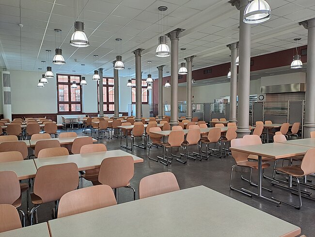 Ein leerer Speisesaal mit vielen Tischen und Stühlen, die in Reihen angeordnet sind. Große Fenster lassen Licht herein, und die Decke hat mehrere Lampen. Im Hintergrund befindet sich eine Küchenzeile mit Geräten. Der Raum wirkt sauber und ordentlich.