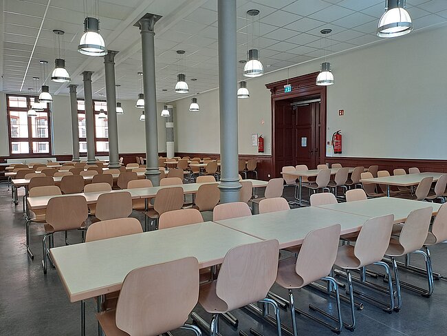 Ein leerer Speisesaal mit mehreren Tischen und Stühlen. Die Tische sind in Reihen angeordnet und haben weiße Tischplatten. Die Stühle sind aus Holz mit Metallbeinen. An der Decke hängen mehrere Lampen, und es gibt große Fenster an einer Wand.