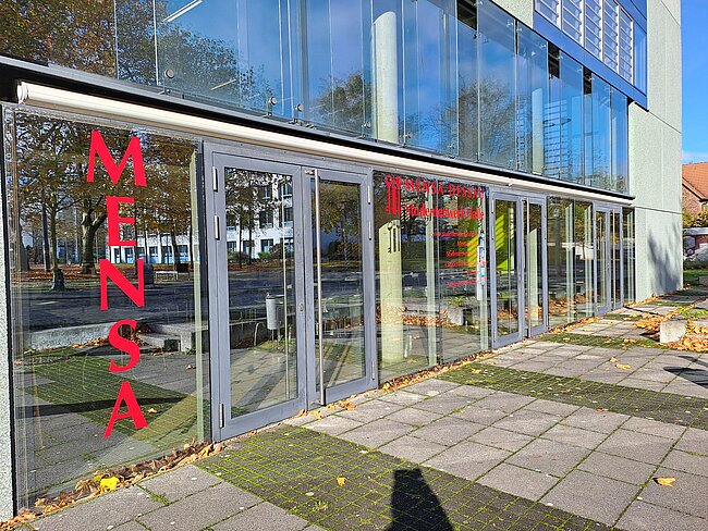 "Ein modernes Gebäude mit großen Glasfenstern. Auf den Fenstern steht in roter Schrift 'Mensa'. Im Hintergrund sind Bäume und ein Gehweg zu sehen. Die Mensa scheint ein Teil einer Bildungseinrichtung zu sein."
