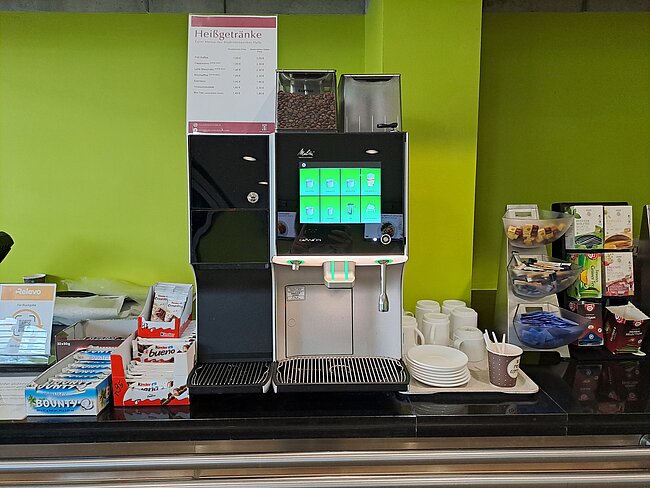 Ein Selbstbedienungs-Kaffeeautomat mit Touchscreen-Bedienfeld, daneben stehen Tassen und Untertassen bereit. Auf dem Tresen befinden sich verschiedene Snacks und Süßigkeiten. Ein Schild mit Getränkepreisen ist über dem Automaten angebracht.