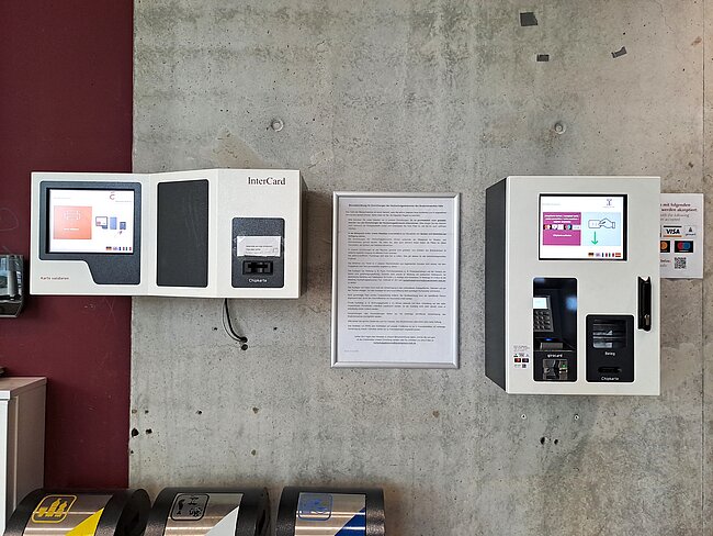"Zwei InterCard-Geräte an einer Wand mit einem Informationsblatt dazwischen. Die Geräte dienen zur Identifikation und haben Touchscreens sowie Kartenleser. Das Informationsblatt enthält Anweisungen oder Erklärungen zur Nutzung der Geräte."