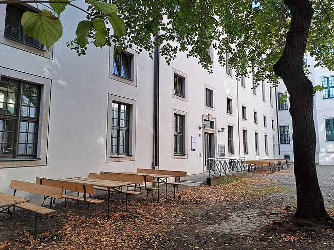 Ein weißes Gebäude mit mehreren Fenstern und Tischen mit Bänken davor. Die Tische und Bänke sind aus Holz und stehen auf einem gepflasterten Bereich mit Laub auf dem Boden. Ein großer Baum steht neben den Tischen und spendet Schatten.