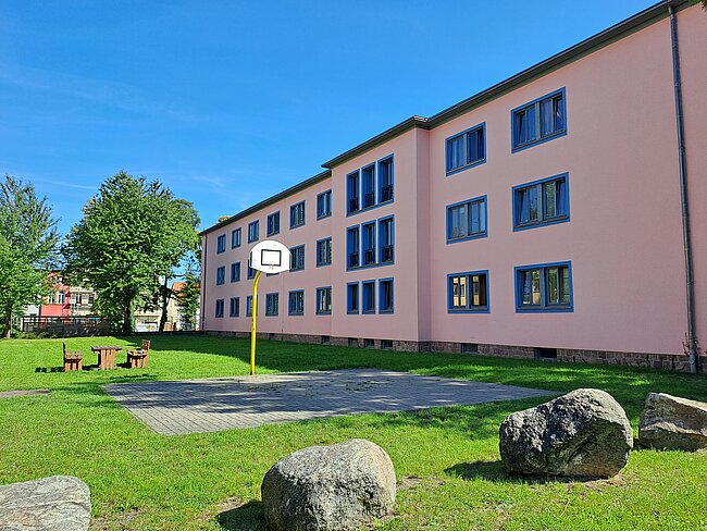 Ein pinkfarbenes Gebäude mit mehreren Fenstern, davor ein Basketballkorb auf einem Betonplatz. Im Vordergrund befinden sich große Steine und ein Bereich mit Rasen. Links stehen ein paar Bänke und ein Baum. Der Himmel ist klar und blau.