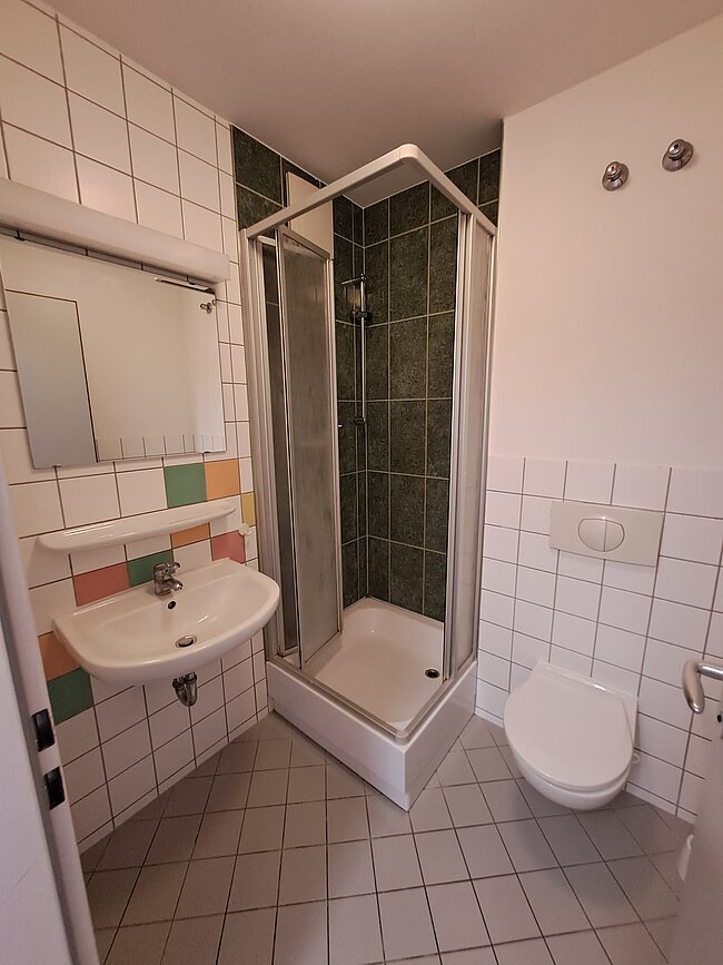 Ein kleines Badezimmer mit Waschbecken, Toilette und Duschkabine. Die Wände sind mit weißen Fliesen und farbigen Bordüren verkleidet. Die Duschkabine hat grüne Fliesen und eine klare Glasscheibe. Ein Handtuchhalter ist über dem Waschbecken angebracht.