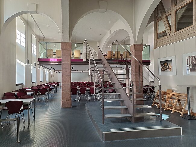 Ein moderner Raum mit mehreren Tischen und Stühlen, einer Treppe mit Metallgeländer und großen Fenstern. Die Wände sind weiß mit hohen Bögen, und es gibt eine Galerie im ersten Stock mit weiteren Sitzgelegenheiten. Der Boden ist mit blauen Fliesen bedeckt.