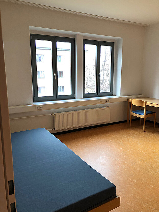 Zimmer WH 2 Köthen; Foto: Thomas Faust Ein einfaches, spartanisch eingerichtetes Zimmer mit einem Bett, einem Schreibtisch mit Stuhl und einem großen Fenster. Das Fenster zeigt eine Aussicht auf andere Gebäude. An der Wand befindet sich ein Heizkörper. Der Boden ist mit Linoleum bedeckt.
