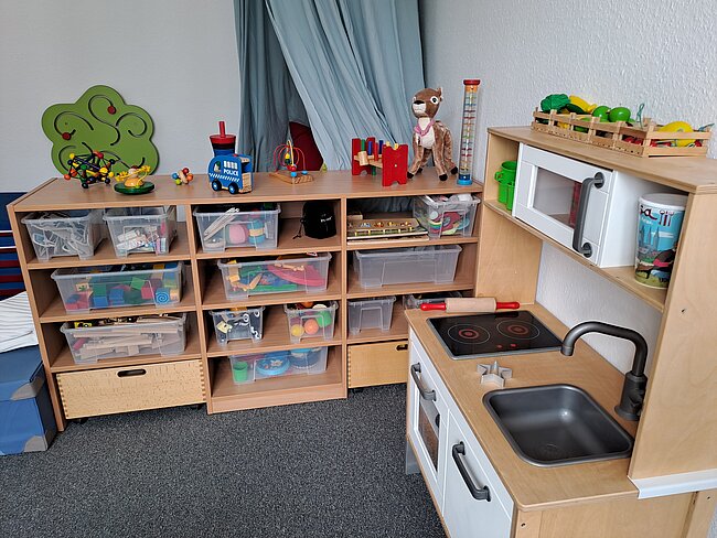 Spielküche mit Spüle, Herdplatte und Wasserhahn, daneben Regal mit durchsichtigen Fächern voller Spielzeug wie Bausteine, Fahrzeuge, Puppen und Alltagsgegenstände sowie einer grünen Baumwand.
