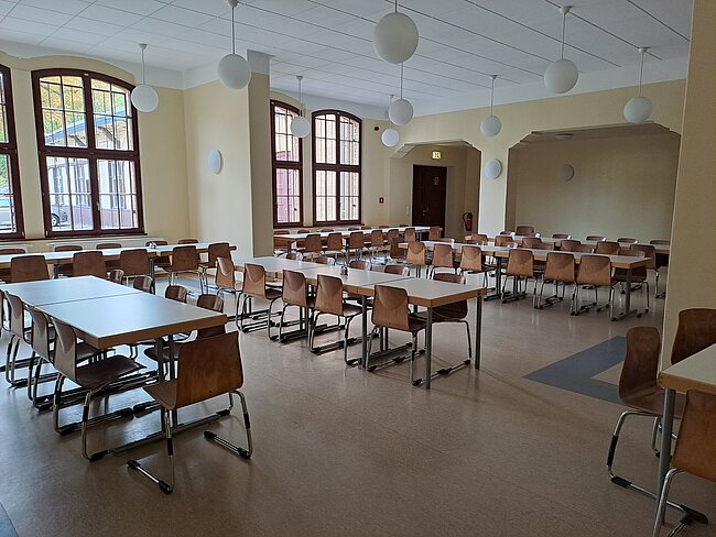 Ein leerer Speisesaal mit mehreren Tischen und Stühlen, großen Fenstern an der linken Wand und runden, weißen Deckenlampen. Der Raum wirkt sauber und ordentlich, mit hellen Wänden und einem glatten Boden. Es gibt eine Tür in der Mitte der Rückwand.