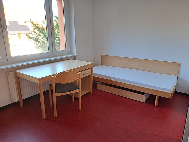 Ein einfaches Zimmer mit einem Schreibtisch, einem Stuhl und einem Bett. Der Schreibtisch hat eine Schublade und steht neben einem Fenster. Das Bett ist ein einfaches Holzgestell mit einer Matratze. Der Boden ist mit rotem Teppich ausgelegt.