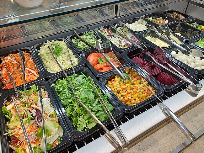 Ein Buffet mit verschiedenen Salaten und Beilagen. Es gibt grüne Salate, Karotten, Bohnen, Kohlrabi, Tomaten, Gurken und andere frische Zutaten. Die Salate sind in schwarzen Schüsseln mit Servierlöffeln angeordnet.