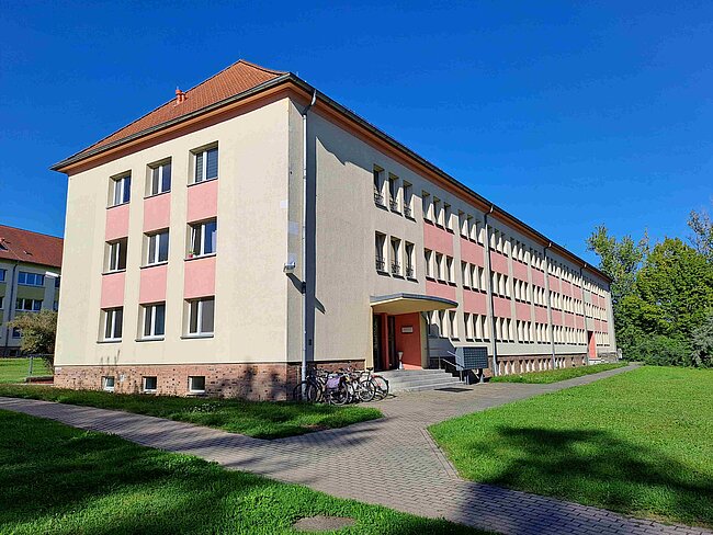 Ein mehrstöckiges Gebäude mit hellbeiger Fassade und roten Akzenten. Es hat mehrere Fenster und einen Eingang mit einer kleinen Überdachung. Vor dem Gebäude befinden sich Fahrräder und ein gepflasterter Weg. Der Hintergrund zeigt einen grünen Rasen und einen klaren blauen Himmel.
