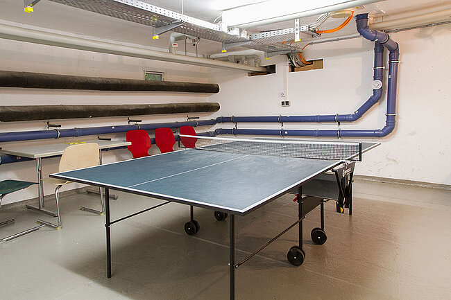 Ein Tischtennistisch steht in einem Raum mit mehreren aufgestapelten Luftkanälen an der Wand. Im Hintergrund sind einige Stühle und ein Tisch zu sehen. Der Raum wirkt wie ein Lager oder ein Technikraum mit einer Deckenkonstruktion aus Metall und Rohren.
