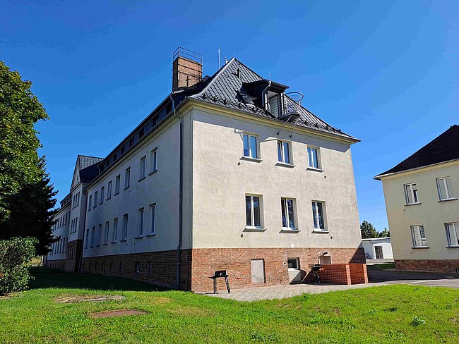 Ein mehrstöckiges Gebäude mit hellgrauer Fassade und braunem Sockel. Es hat mehrere Fenster und ein schräges Dach mit einem Schornstein. Das Gebäude steht auf einem grünen Rasen und ist von Bäumen umgeben. Der Himmel ist klar und blau.