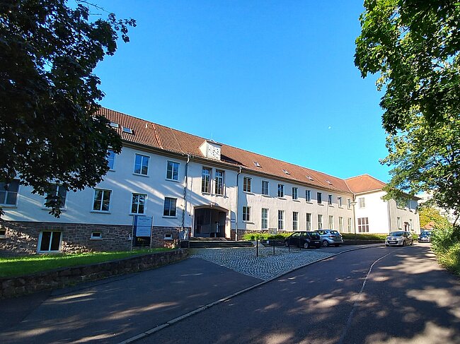 Ein großes, weißes Gebäude mit rotem Dach und mehreren Fenstern. Vor dem Gebäude befindet sich ein gepflasterter Bereich mit einigen geparkten Autos. Der Himmel ist klar und blau, und es gibt Bäume auf beiden Seiten des Gebäudes.