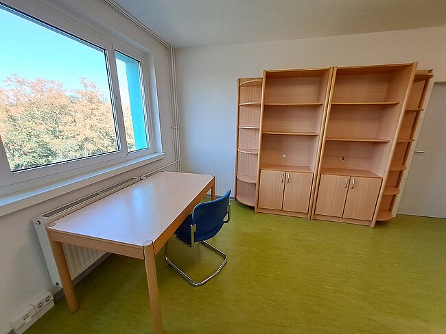 Ein einfacher Raum mit einem Schreibtisch, einem blauen Bürostuhl und einem großen Regal mit Schubladen und Fächern. Ein Fenster mit Blick auf Bäume lässt Tageslicht herein. Der Boden ist in einem hellen Grünton gehalten.
