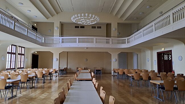 Harzmensa, großer Saal Ein großer, leerer Saal mit vielen Tischen und Stühlen, die in Reihen angeordnet sind. Der Raum hat eine hohe Decke mit einem kreisförmigen Licht an der Decke und eine Galerie im oberen Bereich. Die Wände sind hell und es gibt große Fenster auf der linken Seite.