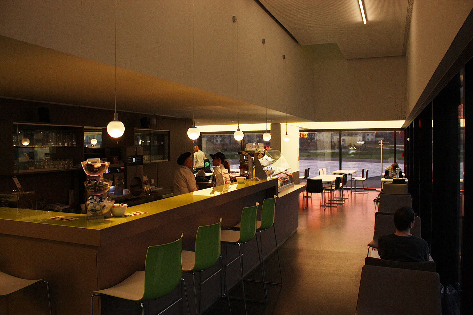 Cafebar Heidemensa; Foto: Thomas Faust Ein Café mit einer Theke, an der zwei Personen stehen. Hinter der Theke sind Regale mit Gläsern und Flaschen. Im Hintergrund sitzen einige Gäste an Tischen. Das Café hat eine moderne, minimalistische Einrichtung mit grünen Stühlen an der Theke und einer großen Fensterfront.