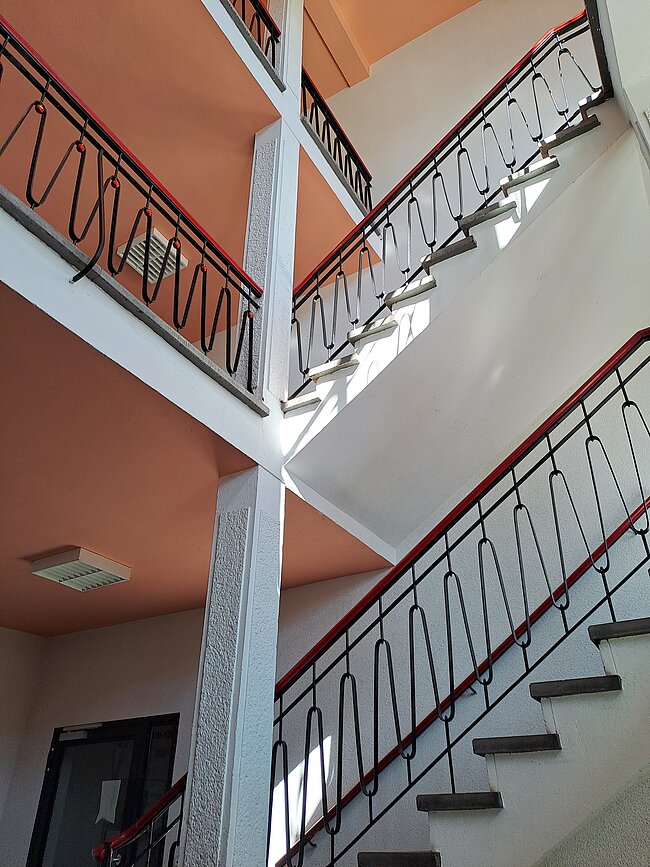 Ein Treppenhaus mit zwei Etagen und schwarzen schmiedeeisernen Geländern. Die Wände sind in hellen Farben gehalten, und natürliches Licht fällt durch Fenster herein. Die Treppe hat weiße Stufen mit roten Kanten.
