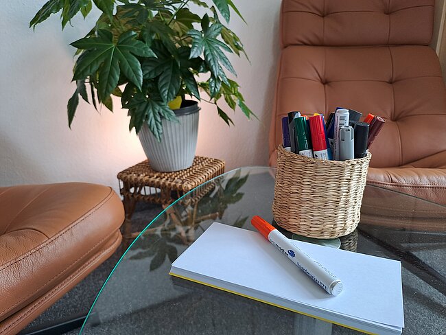 Auf einem Glastisch liegen Notizzettel und ein weißer Stift darauf. Daneben steht ein Korb mit bunten Stiften und Markern. Im Hintergrund ist eine grüne Zimmerpflanze auf einem Hocker zu sehen.
