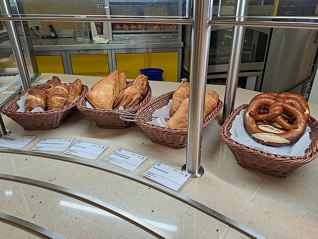 "Ein Buffet mit verschiedenen Brotsorten und Brezeln in Körben. Die Brote sind in unterschiedlichen Formen und Größen. Die Körbe stehen auf einem runden Tisch mit Beschilderung. Im Hintergrund ist eine Küche mit gelben Wänden zu sehen."
