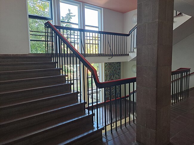 Ein Treppenhaus mit einem Holzgeländer, das rot gestrichen ist. Die Treppe führt nach oben zu einem oberen Stockwerk mit großen Fenstern, die Tageslicht hereinlassen. Die Wände sind teilweise mit Fliesen verkleidet.