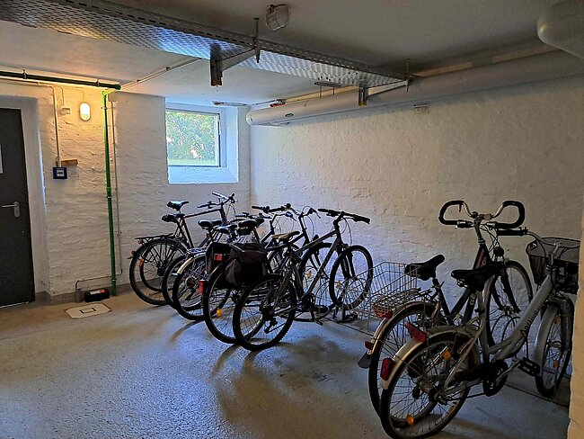 "Ein Fahrradkeller mit mehreren Fahrrädern, die an der Wand befestigt sind. Der Raum hat eine weiße, raue Wand und einen grauen Boden. Ein kleines Fenster lässt etwas Licht herein. Die Fahrräder sind in einer Reihe aufgestellt und teilweise mit Körben oder Taschen ausgestattet."