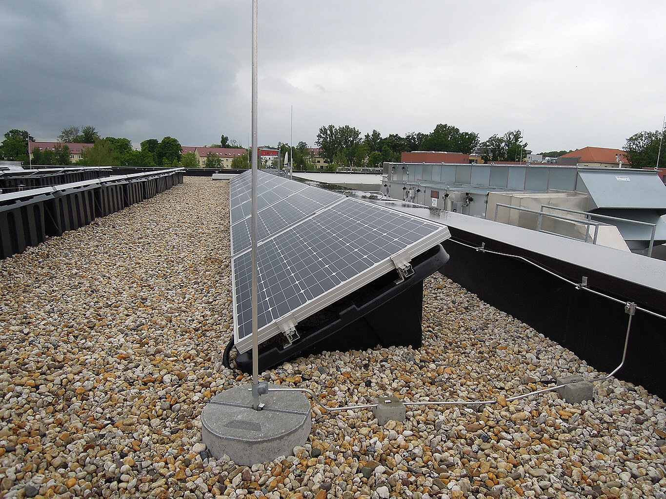 Dach Heidemensa; Foto: Thomas Faust "Ein Solarmodul auf einem mit Kies bedeckten Dach. Im Hintergrund sind weitere Solarmodule und Gebäude zu sehen. Der Himmel ist bewölkt."
