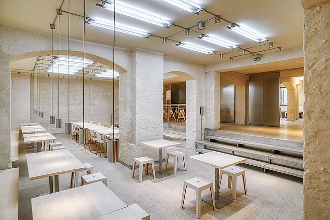 Speisesaal (© Patrick Müßiggang) Ein moderner, minimalistischer Raum mit hellen Wänden und Boden. Es gibt mehrere Tische mit Hockern und Bänken. Der Raum ist durch Bögen unterteilt und hat eine offene Treppe. Die Beleuchtung ist hell und gleichmäßig, was eine klare und ruhige Atmosphäre schafft.