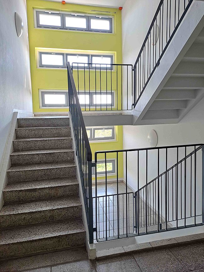 Ein Treppenhaus mit zwei Treppenläufen und einem Geländer. Die Wände sind weiß und gelb gestrichen, mit mehreren Fenstern, die natürliches Licht hereinlassen. Der Boden und die Stufen sind aus grauem Stein. Die Treppen führen zu verschiedenen Stockwerken.