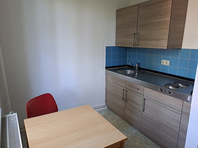 Ein kleiner Raum mit einem Tisch und einem Stuhl. An der Wand befindet sich eine Küchenzeile mit Arbeitsplatte, Spüle, zwei Herdplatten und Schränken darüber. Die Wände sind weiß und blau gefliest. Der Boden ist mit Teppich ausgelegt.