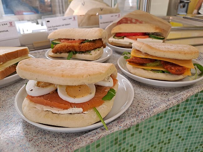 "Mehrere belegte Brötchen auf Tellern. Die Brötchen enthalten verschiedene Zutaten wie Eier, Salat, Tomaten und Fleisch. Die Brötchen sind auf einem Tisch in einer Cafeteria oder einem Imbiss angeordnet."