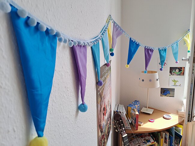 Bunte Girlande mit dreieckigen, pastellfarbenen Bändern in Blau, Violett und Pink an einer Wand aufgehängt, verziert mit kleinen Pompons und Glöckchen. Im Hintergrund ein Kinderzimmer mit Regal, Büchern und Spielzeug.