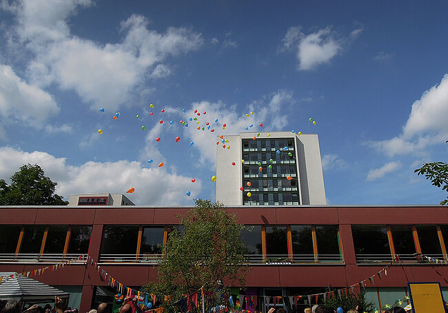"Ein Gebäude mit mehreren Stockwerken im Hintergrund, davor eine Menschenmenge. Bunte Luftballons steigen in den blauen Himmel mit einigen Wolken auf. Die Atmosphäre wirkt festlich und fröhlich."